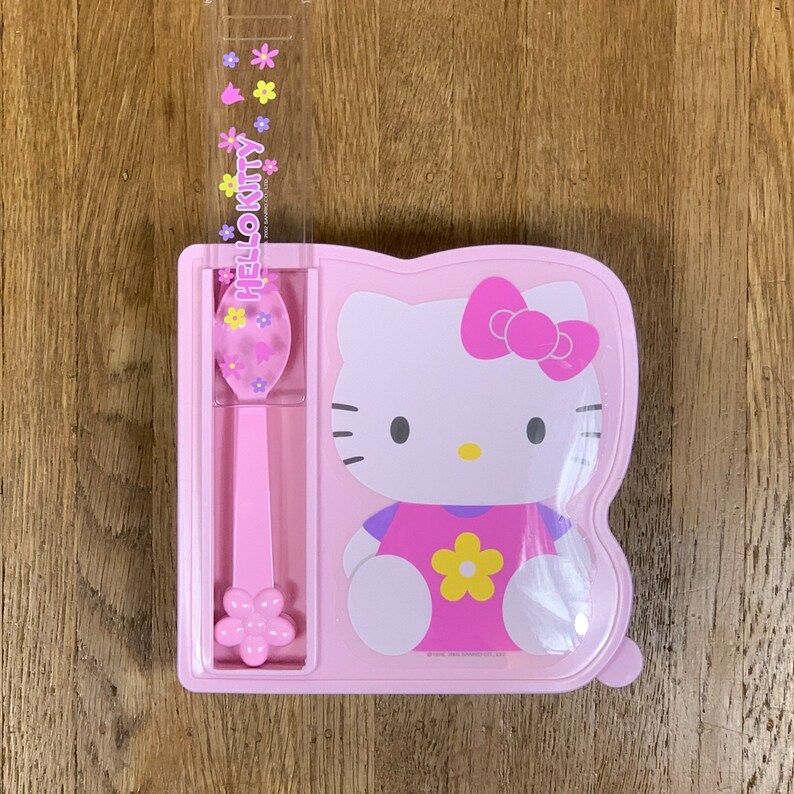 May include: Pink Hello Kitty lunchbox with a matching pink spoon. The lunchbox has a picture of Hello Kitty on the front and a compartment for the spoon.