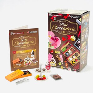 May include: A miniature chocolate shop set with a box, brochure, and various chocolate pieces. The box and brochure feature the text "Petit Chocolaterie." The set includes a three-tiered serving tray, small boxes, and tongs. The color scheme is brown, red, and orange.