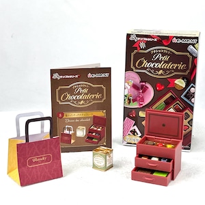 May include: Miniature chocolate shop set featuring a red three-drawer chest, a box, a brochure, a shopping bag, and a small tin. The box and brochure read "Petit Chocolaterie". The set includes various chocolate-themed accessories.