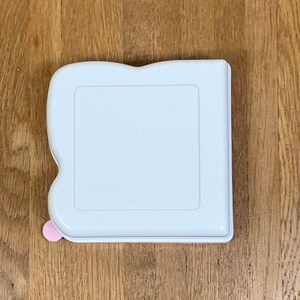May include: White plastic storage container with a rounded edge and a pink button on the side.