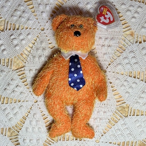Puede incluir: Un oso de peluche naranja con una camisa blanca de cuello y una corbata azul y blanca de lunares. El oso está sentado en un encaje blanco de ganchillo.