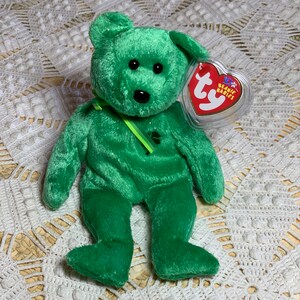 May include: Green plush beanie baby bear with a green shamrock on its chest and a green ribbon around its neck. The bear has black eyes and a black nose. The bear is sitting on a white crocheted surface. The bear has a Ty Beanie Babies tag attached to its ear.