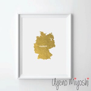 Può includere: Una stampa incorniciata con una mappa dorata della Germania su sfondo bianco. La parola "GERMANY" è stampata in bianco sulla mappa, con "BERLIN" e un piccolo punto che indica la posizione della città. La stampa è in una cornice bianca.