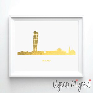 May include: Gold foil print of the Malmö skyline, featuring a tall, modern building and other structures. The print is framed in a white frame and includes the text "MALMÖ" at the bottom.