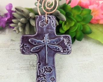 Puede incluir: Cruz de cerámica morada con un diseño de libélula blanca y detalles florales. La cruz tiene un colgador de alambre de cobre.