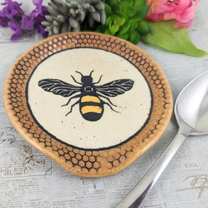 May include: A ceramic spoon rest with a honeycomb design and a black and yellow bee illustration. The spoon rest is brown and beige.