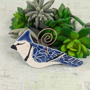 May include: A blue and white ceramic blue jay bird figurine with a copper wire stand. The bird has a detailed floral pattern on its wings and body.