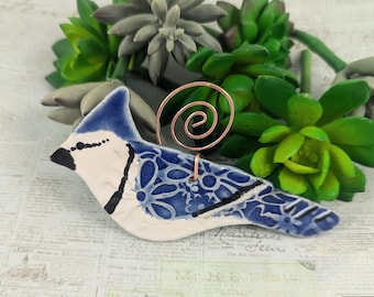 Puede incluir: Una figurita de cerámica azul y blanca de un arrendajo azul con un soporte de alambre de cobre. El pájaro tiene un patrón floral detallado en sus alas y cuerpo.