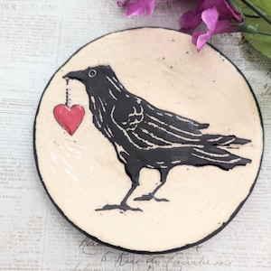 May include: A ceramic dish with a black crow carrying a red heart. The dish has a cream background and black rim.