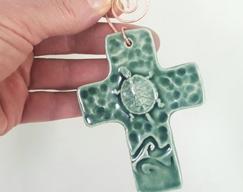 May include: A green ceramic cross with a turtle design and a copper wire hanger. The cross has a textured surface and a wave design at the bottom.