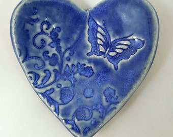 Puede incluir: Un plato de cerámica azul en forma de corazón con una mariposa blanca y un diseño floral.