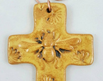 May include: A handmade, ceramic cross pendant with a bee and floral design. The cross is a warm yellow color with a copper wire spiral at the top for hanging. The bee is in the center of the cross.