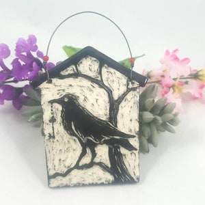 May include: A black and white ceramic ornament depicting a crow perched on a branch. The ornament has a hanging loop and is shaped like a small house.