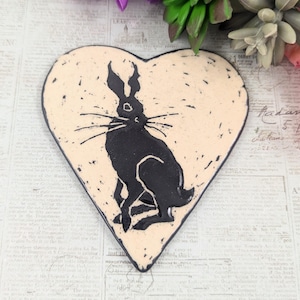 May include: A heart-shaped ceramic piece with a black rabbit silhouette on a cream background. The rabbit is sitting upright with long ears and whiskers. The piece has a rustic, handmade appearance.
