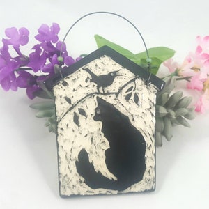 May include: A black and white ceramic house-shaped ornament with a bird and cat silhouette. The ornament has a wire hanger for hanging.