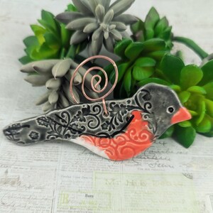 May include: A ceramic bird ornament with a gray and black floral pattern and a red and white belly. The bird has a copper wire loop for hanging.