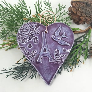 May include: Purple heart-shaped ceramic ornament with a raised design of the Eiffel Tower, a bird, and floral patterns.