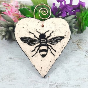 May include: White ceramic heart-shaped ornament with a black and white bee illustration. The ornament has a copper wire hanger.