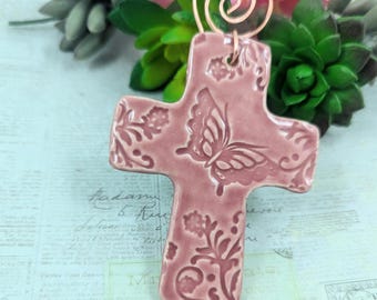 May include: A dusty rose ceramic cross with a butterfly and floral design. The cross has a copper wire spiral at the top. The cross is a decorative item.
