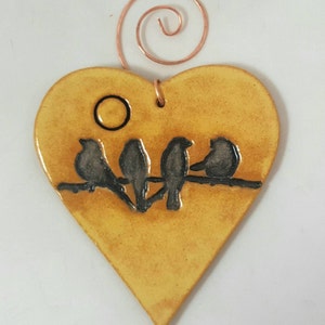 May include: A yellow heart-shaped ceramic ornament with a wire hanger. The ornament features a silhouette of four birds perched on a branch with a small circle representing the sun.