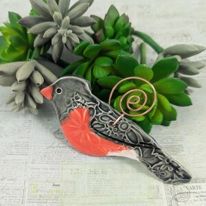 May include: A ceramic bird figurine featuring a dark gray body with a floral pattern, a bright orange chest, and a small orange beak. A copper-colored wire spiral is attached to the back of the bird, extending upward from the ceramic surface.