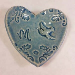 May include: A blue ceramic heart-shaped dish with a white hummingbird design and the letter "M" in the center.
