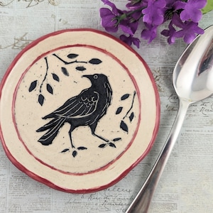May include: A round, off-white ceramic plate with a red rim and a black crow design. The crow is walking on a branch with leaves. A silver spoon and purple flowers are in the background.