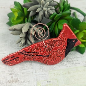 May include: Red cardinal bird ornament with black accents and a floral pattern. The ornament is made of ceramic and has a copper wire hanger.