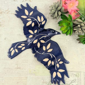 May include: A navy blue ceramic raven wall hanging with cream-colored leaf accents. The raven is depicted in flight, with outstretched wings. Decorative art piece for home decor.