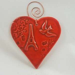 May include: A heart-shaped, red ceramic ornament with a copper wire hanger. The ornament features embossed designs of the Eiffel Tower, a bird, and floral patterns. The heart measures approximately 3 inches wide.