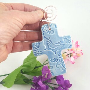 May include: A light blue ceramic cross with a butterfly design and a copper wire hanger. The cross is decorated with a floral pattern.