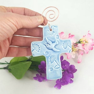 May include: A light blue ceramic cross with a hummingbird design and floral accents. The cross has a copper wire hanger.