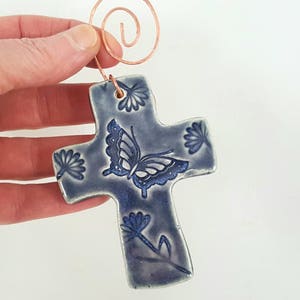 Puede incluir: Una cruz de cer&aacute;mica azul con un dise&ntilde;o de mariposa y flores. La cruz tiene un colgador de alambre de cobre.