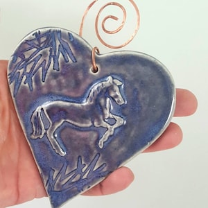 May include: A heart-shaped ceramic ornament with a glossy blue and purple glaze. The surface features a raised, light-colored relief of a horse in motion, surrounded by stylized tree branches. A copper wire spiral is attached to the top.