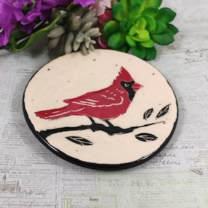 May include: A ceramic plate with a hand-painted red cardinal perched on a black branch with leaves. The plate has a white background with a black rim.