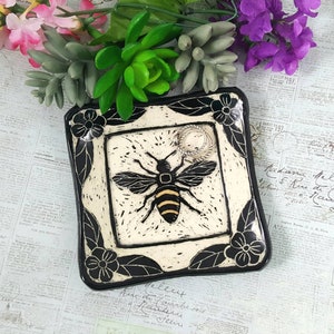 May include: A white ceramic ring dish with a black bee design and black floral accents. The dish has a square shape with rounded corners and a raised rim. A silver ring is resting on the dish.
