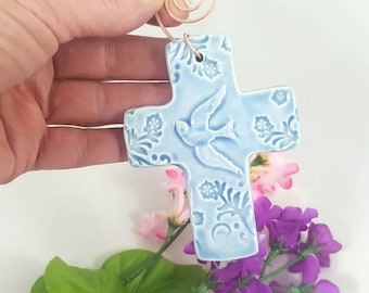 May include: A light blue ceramic cross with a white dove and floral design. The cross has a copper wire hanger.