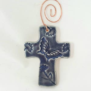 Puede incluir: Un adorno de cruz de cer&aacute;mica azul con un dise&ntilde;o de mariposa y flores. La cruz tiene un colgador de alambre de cobre.