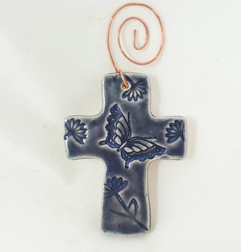 Puede incluir: Un adorno de cruz de cer&aacute;mica azul con un dise&ntilde;o de mariposa y flores. La cruz tiene un colgador de alambre de cobre.