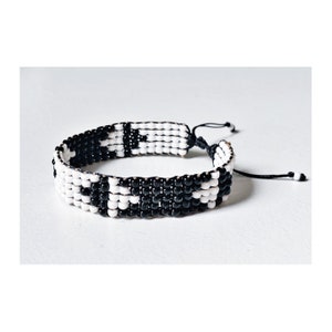 May include: A woven bracelet featuring a repeating pattern of black and white beads. The bracelet has a sliding knot closure for adjustable sizing. The design is a bold, graphic statement piece, suitable for everyday wear.