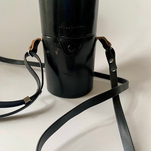 May include: Black leather camera lens case with a strap. The case has a metal clasp and a brown leather strap.