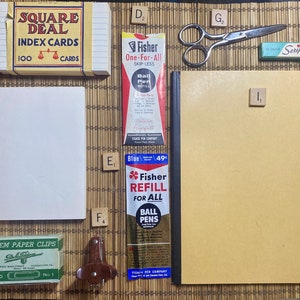 May include: A flat lay of various office supplies on a bamboo mat. The items include a pack of index cards labeled "Square Deal", a pack of paper clips, a small hole punch, a pack of ballpoint pen refills, a notebook, a pair of scissors, and a pack of erasers.