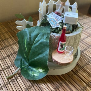 May include: A miniature fairy garden scene with a white picket fence, a small wooden sign, a bottle of red liquid, a glass, and a green leaf. The scene is set in a small wooden pot with moss and a few playing cards.