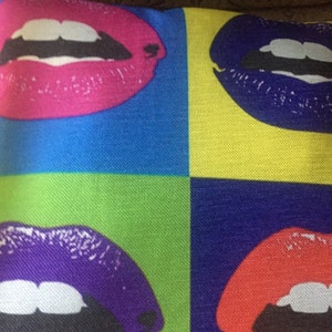 Puede incluir: Una almohada con un diseño pop art que presenta cuatro labios coloridos. Los labios están en un patrón repetitivo de rosa, morado, azul y rojo.