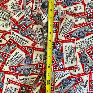 May include: Close-up of white denim pants with a repeating pattern of red, white, and blue Budweiser beer labels. The pants are measured at approximately 14 inches in length. The button and zipper are visible.