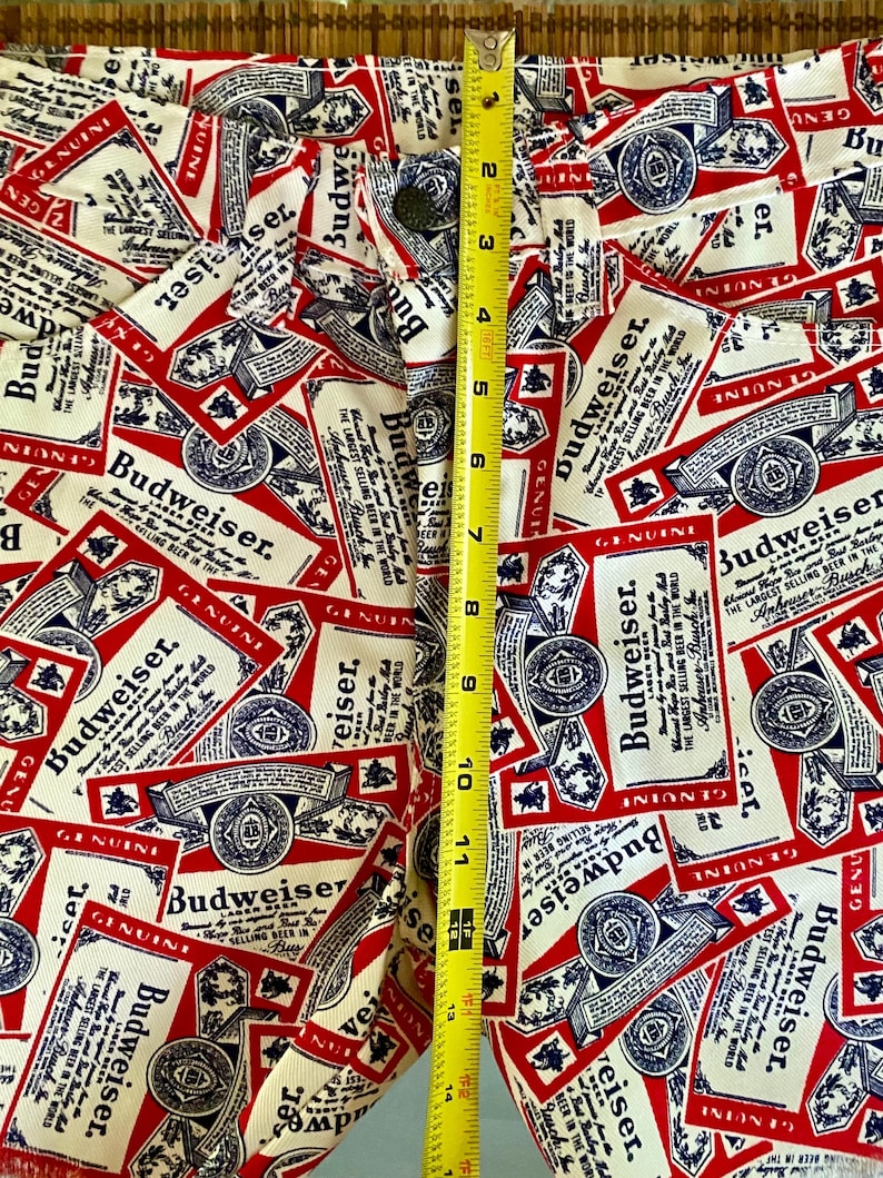 May include: Close-up of white denim pants with a repeating pattern of red, white, and blue Budweiser beer labels. The pants are measured at approximately 14 inches in length. The button and zipper are visible.