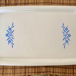 May include: White rectangular serving tray with rounded edges. The tray features a blue floral design on each side. The tray is sitting on a brown bamboo mat. A vintage serving piece for the kitchen.