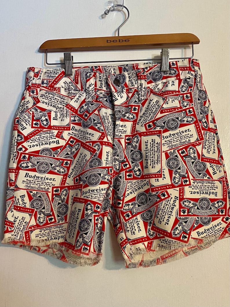 May include: White and red Budweiser-themed shorts with a repeating label pattern. The shorts have a button closure and frayed edges. The shorts are hanging on a wooden hanger.