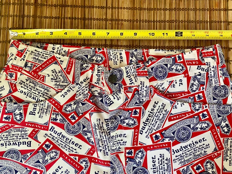 May include: Close-up of white fabric with a repeating pattern of red and blue Budweiser beer labels. The labels feature the Budweiser logo and text. The fabric is likely part of a pair of shorts or pants.