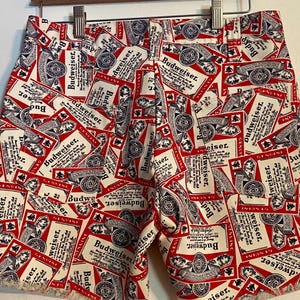 May include: Red and white shorts with a repeating Budweiser label pattern. The shorts are made of a light fabric and have a frayed hem. They are hanging on a wooden hanger.